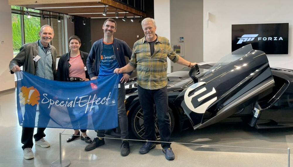 Four smiling people holding a flag in front of a racing car.