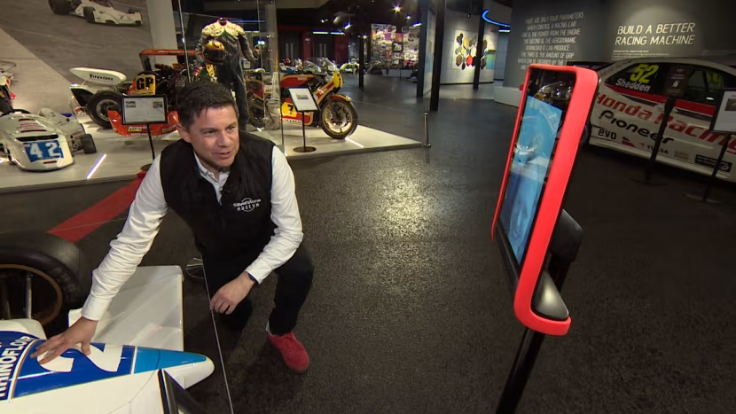 Man crouching in front of racing car talking to wheeled tablet robot